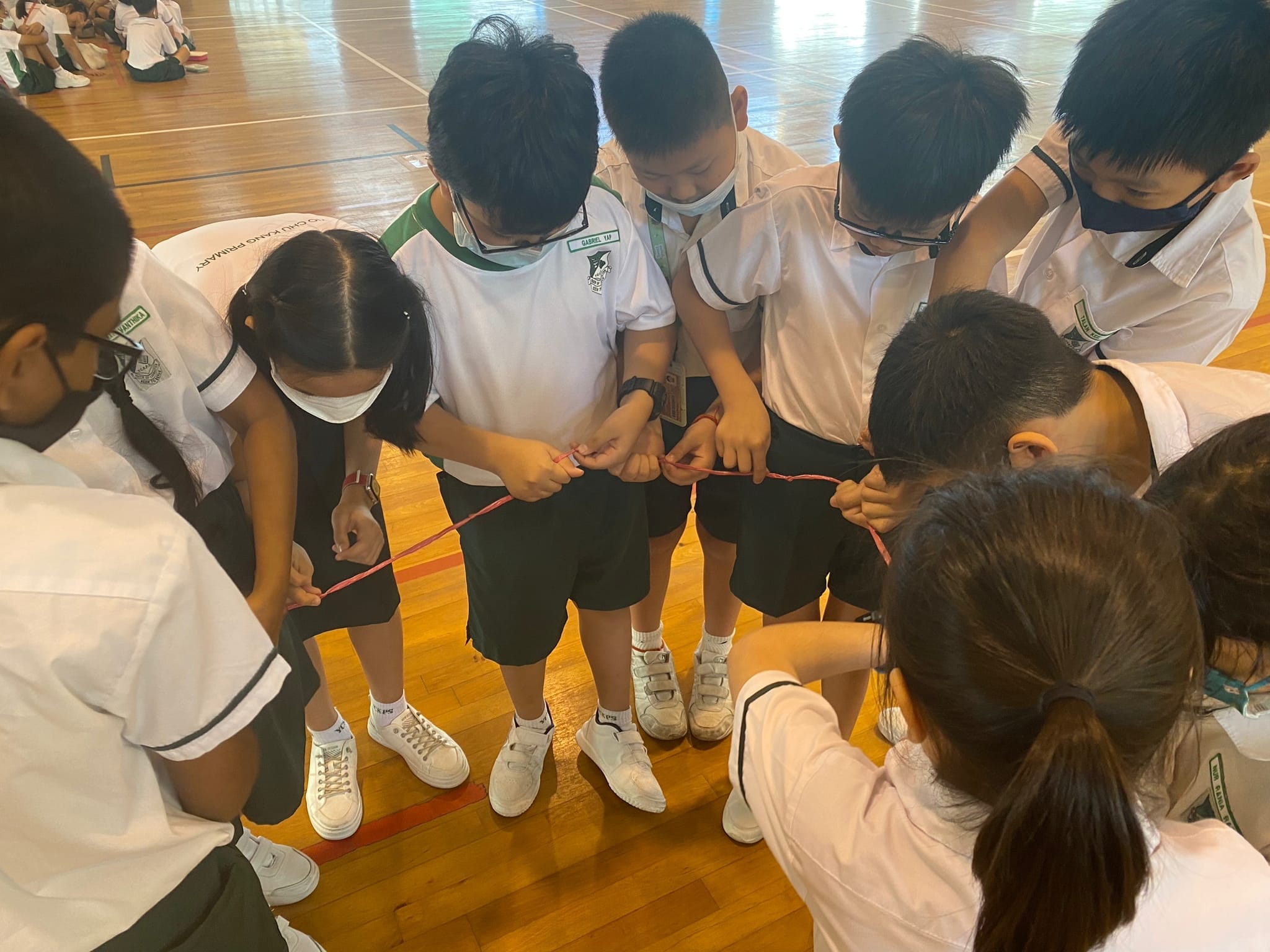 A group of uniformed primary school students wearing masks huddle together, holding a red string in a gym.
